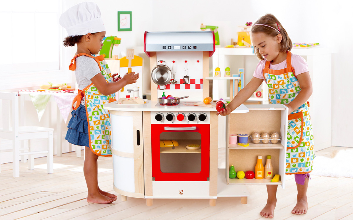 Zwei Kinder spielen mit der Küchentraum-Spielküche von Hape und viel Zubehör. Die Küche ist aus Holz mit vielen Details wie einer roten Backofentür und einer Dunstabzugshaube. Die beiden Mädchen tragen Schürzen und eine Kochmütze beim Fantasie-Rollenspiel.