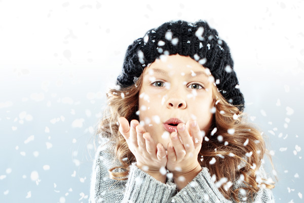 Ein Mädchen mit Wollmütze spielt im Schnee und bläst Schneeflocken aus ihren Händen.